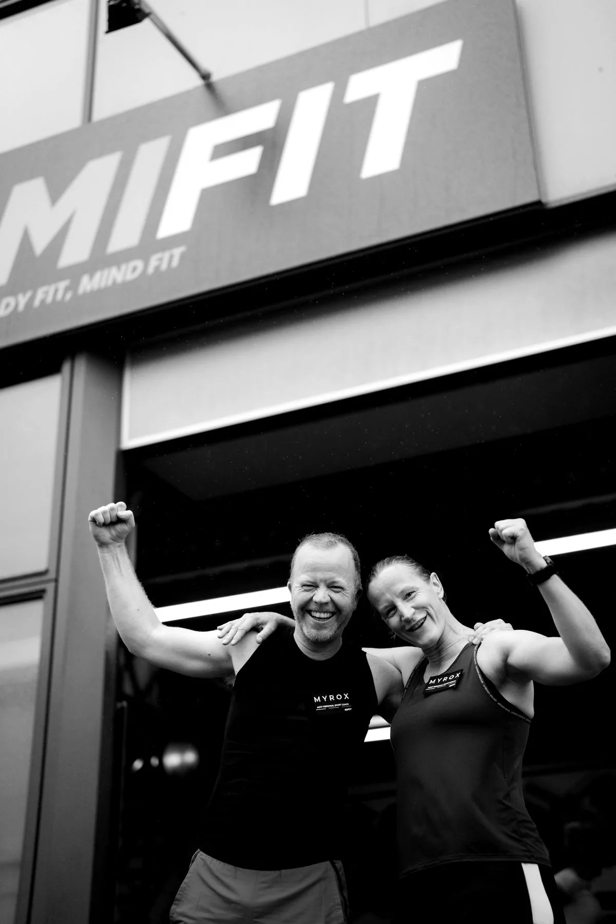 Twee leden juichen met de armen omhoog voor de gevel van Mifit