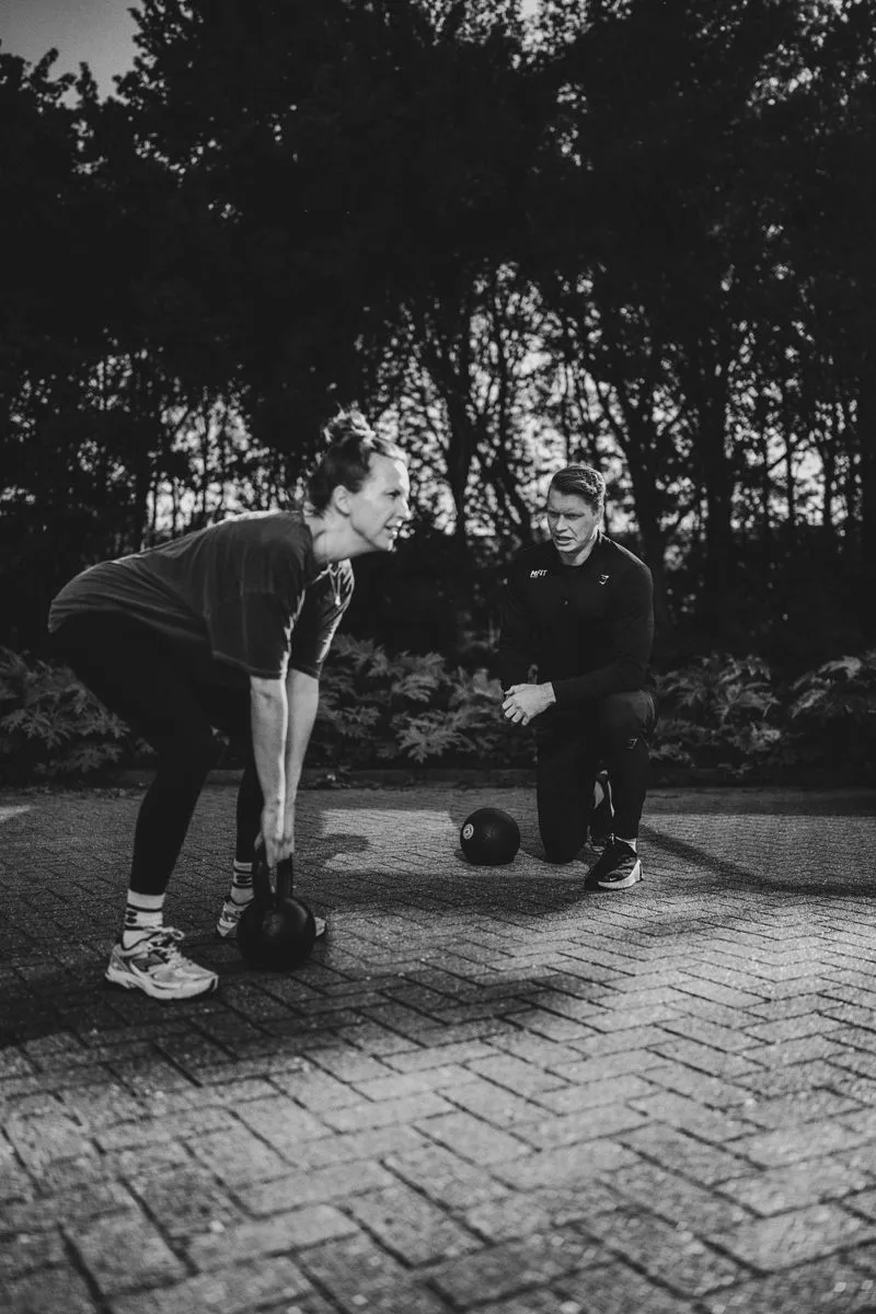 Een personal trainer kijkt geknield mee naar de techniek van een klant met een kettlebell